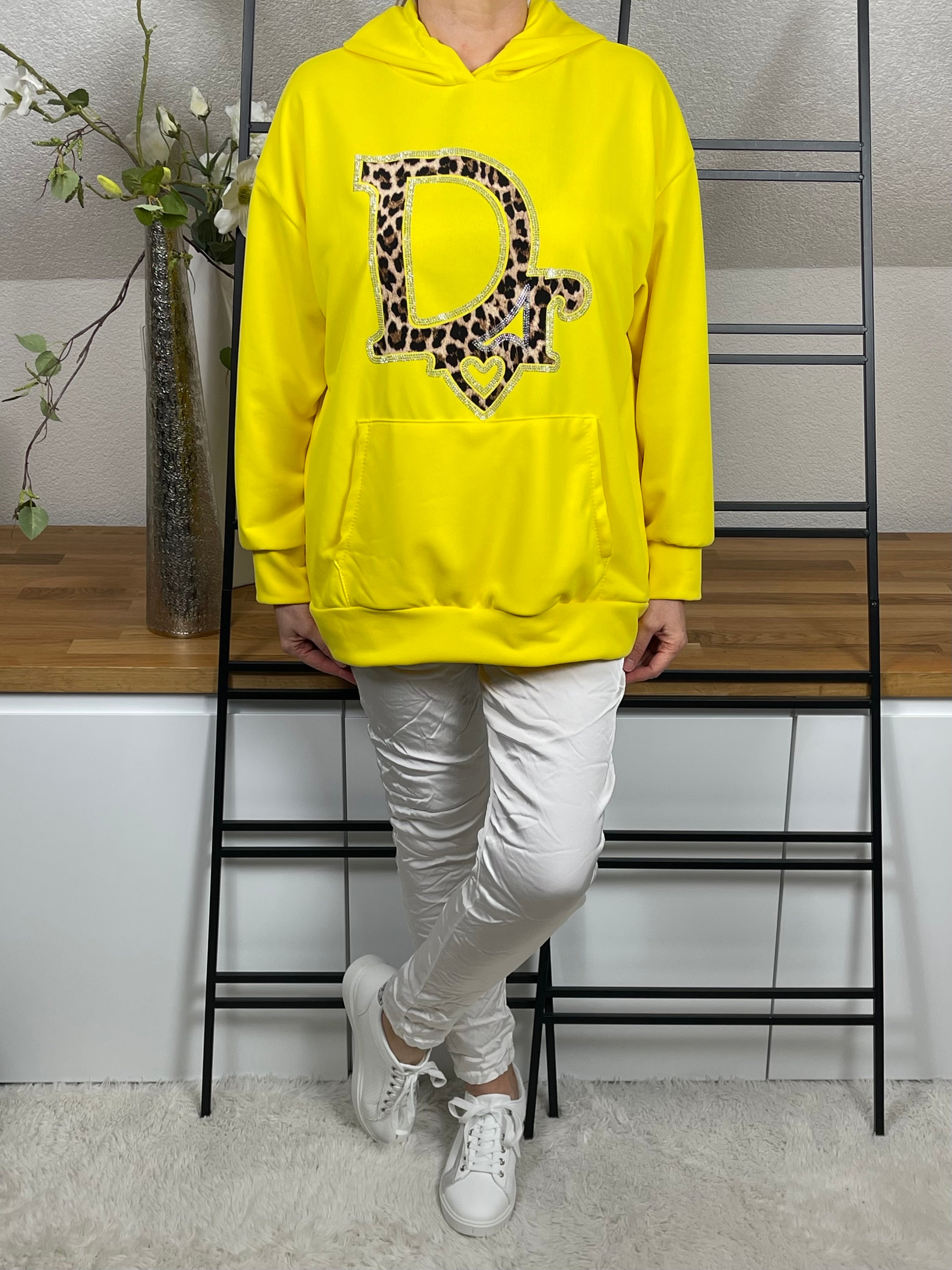Frontansicht eines gelben Hoodies mit Glitzer und Animal-Print, getragen mit gekreuzten Beinen. Die Kordeln des Hoodies sind nach innen geschlagen, kombiniert mit weißer Shiney - Hose und weißen Sneakern
