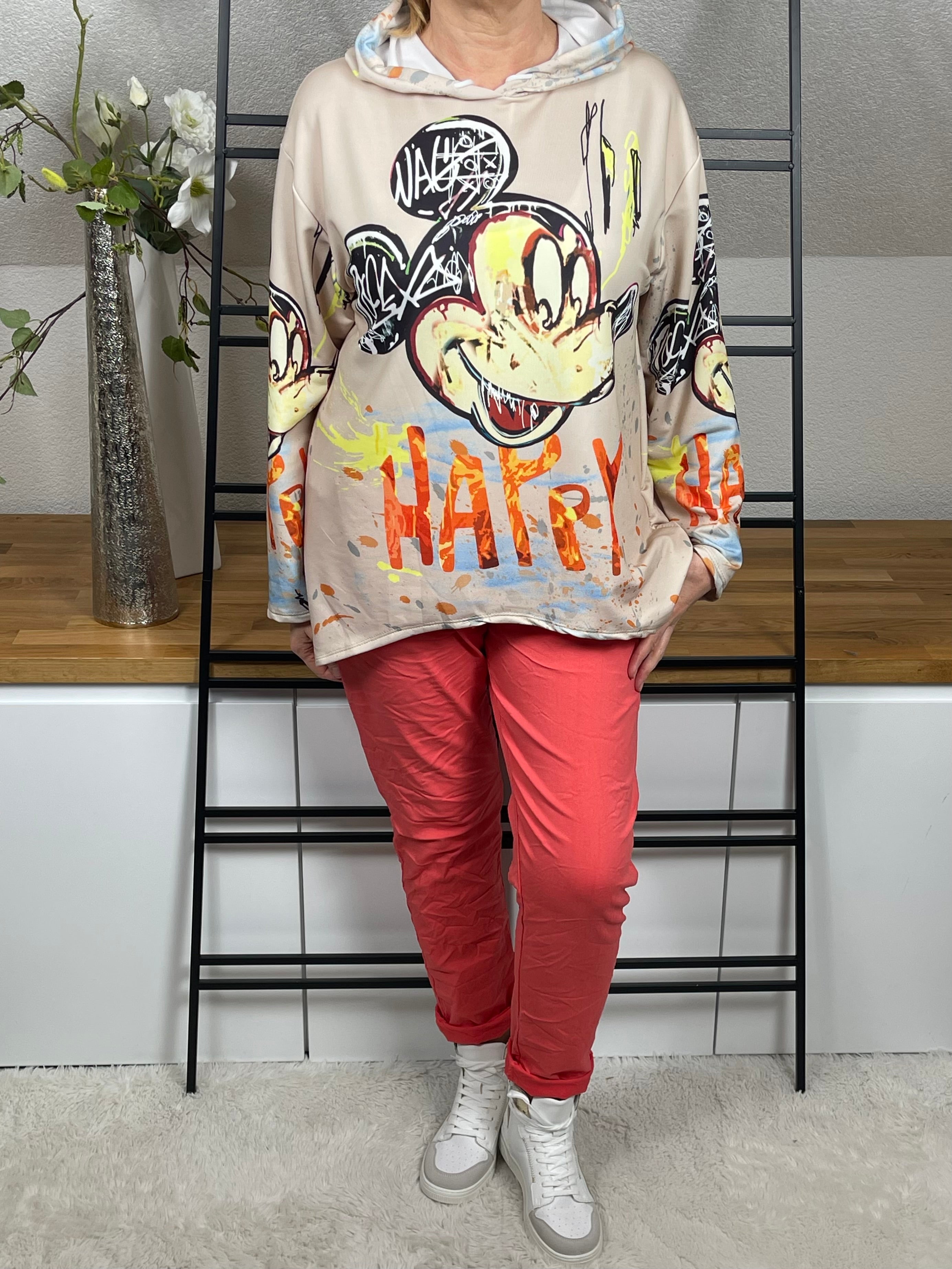 Oversize-Hoodie mit Mickey - Print in beige, in voller Länge, locker geschnitten, kombiniert mit einer korallfarbenen Jogpant und weiß - goldenen High - Sneakern. Perfekt für einen lässigen und bequemen Stil.
