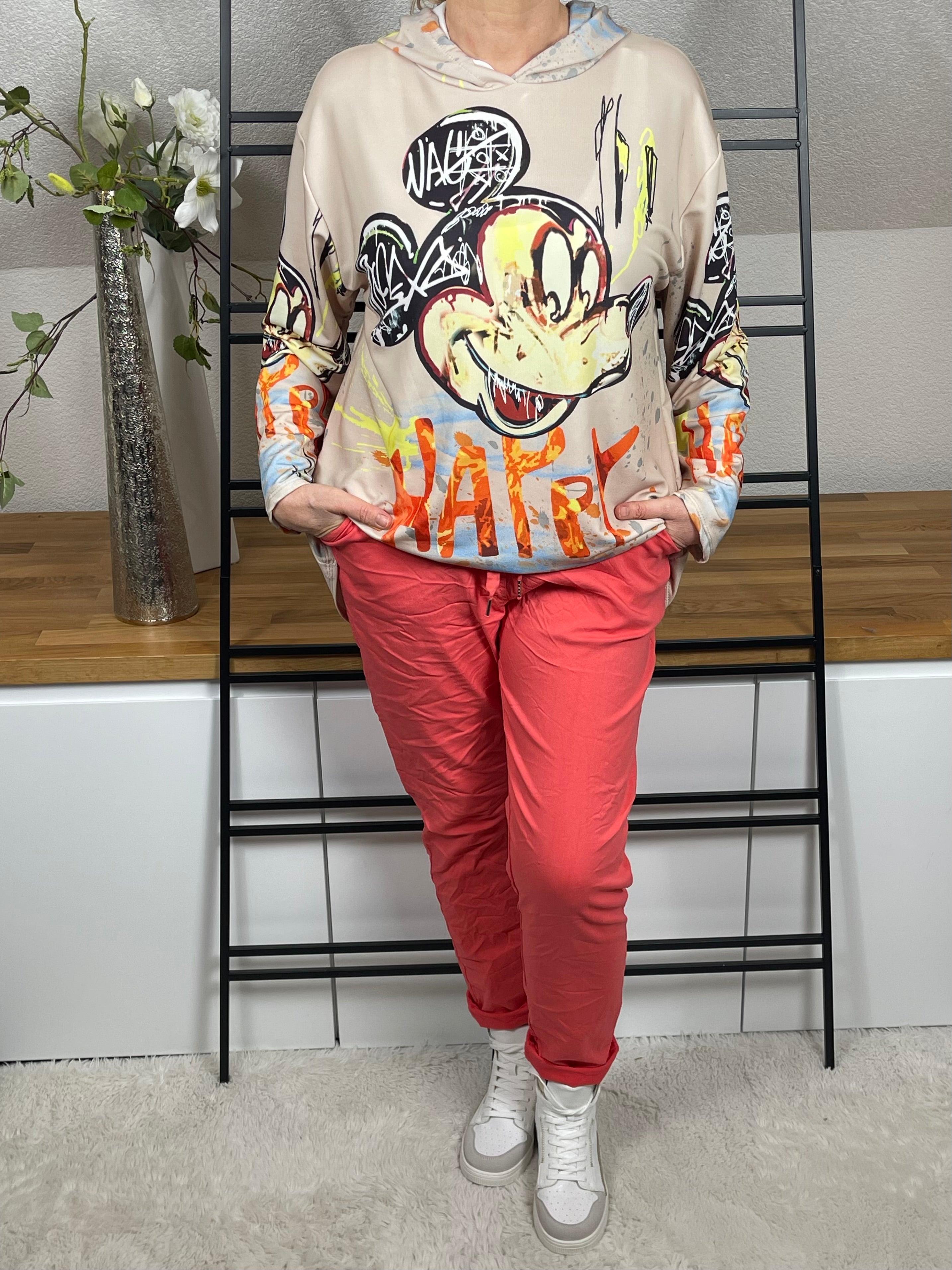 Oversize-Hoodie mit Mickey - Print in beige, vorne leicht nach innen geschlagen getragen, kombiniert mit einer korallfarbenen Jogpant und weiß - goldenen High - Sneakern. Trendiger Look mit bequemem Sitz.