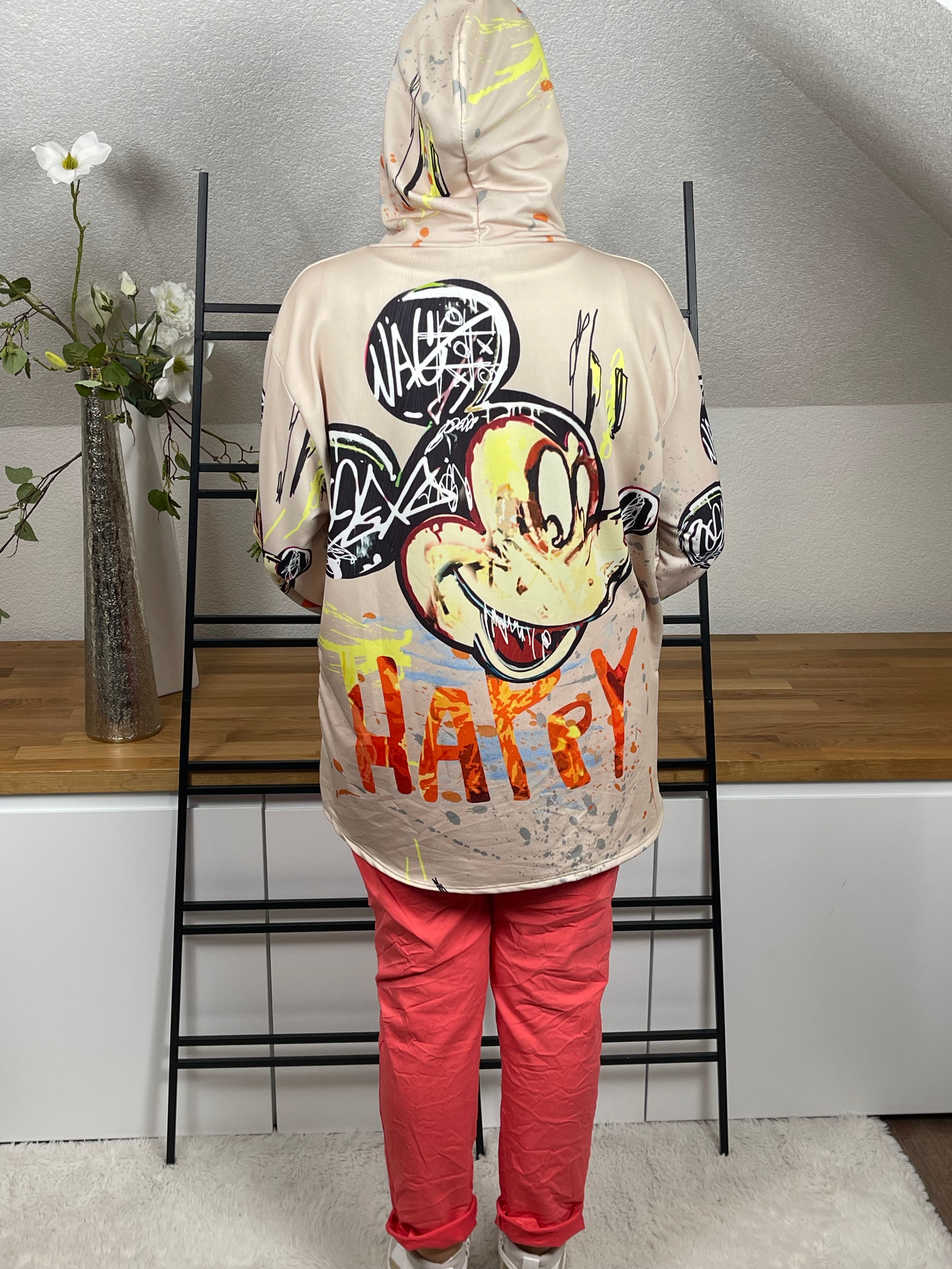 Oversize-Hoodie in beige, mit Mickey - Print von hinten, zeigt die locker fallende Passform und die stylische Kapuze. Kombiniert mit einer korallfarbenen Jogpant und weiß  - goldenen High - Sneakern für einen modernen Look.