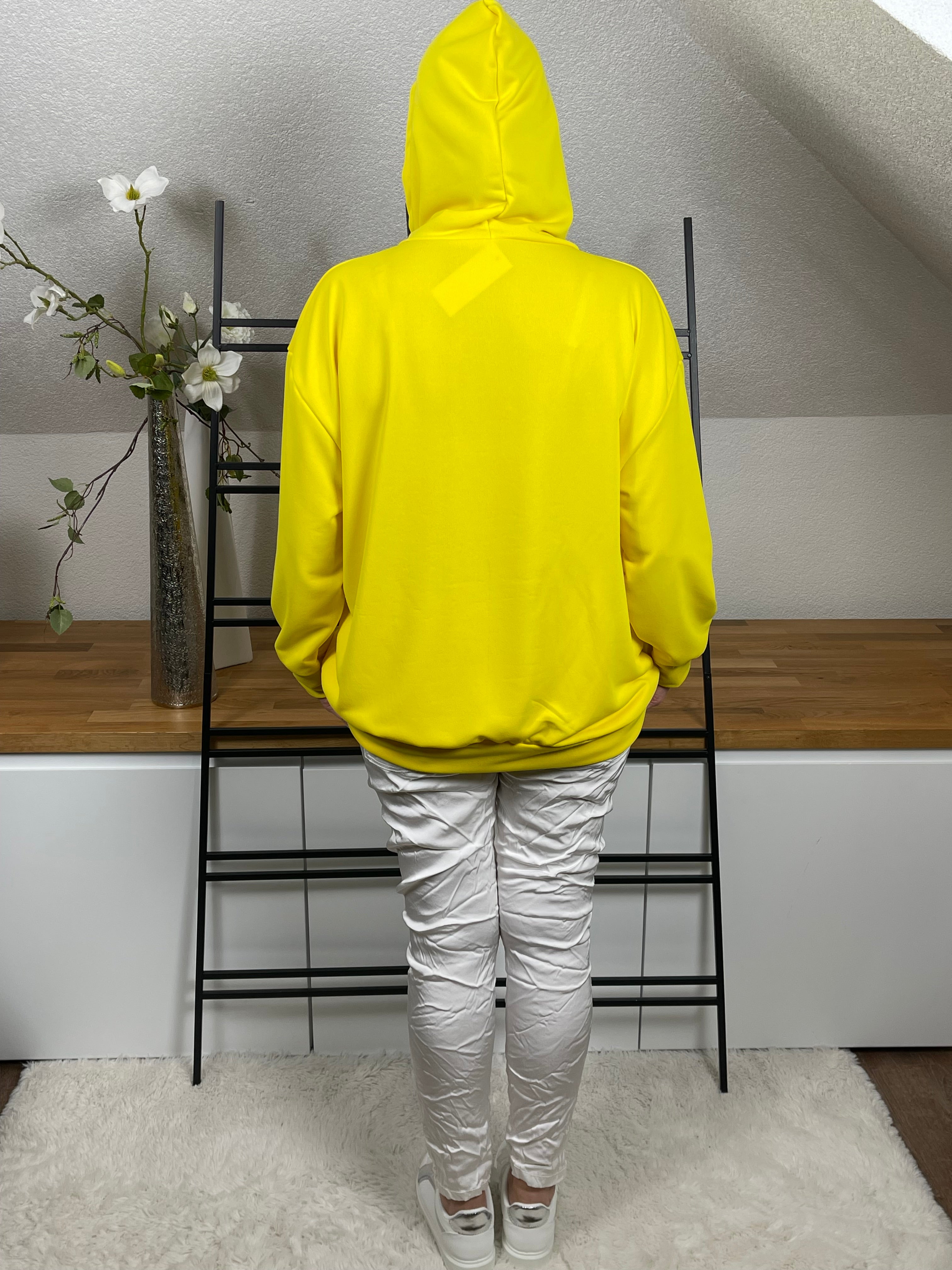 Rückenansicht eines gelben Hoodies mit aufgesetzter Kapuze, kombiniert mit weißer Hose