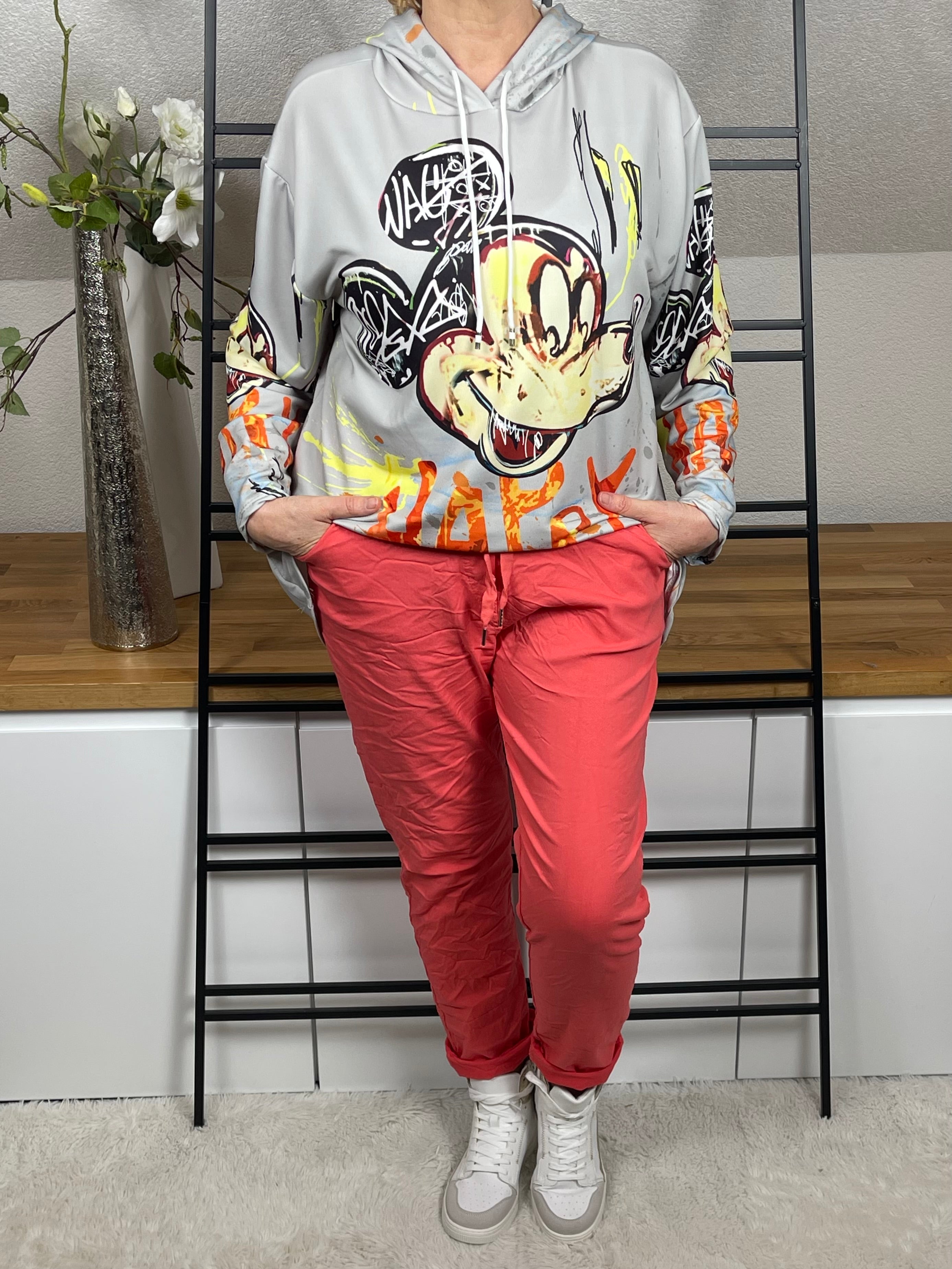 Oversize-Hoodie mit Mickey - Print in hellgrau, vorne leicht nach innen geschlagen getragen, kombiniert mit einer korallfarbenen Jogpant und weißen Sneakern. Trendiger Look mit bequemem Sitz.