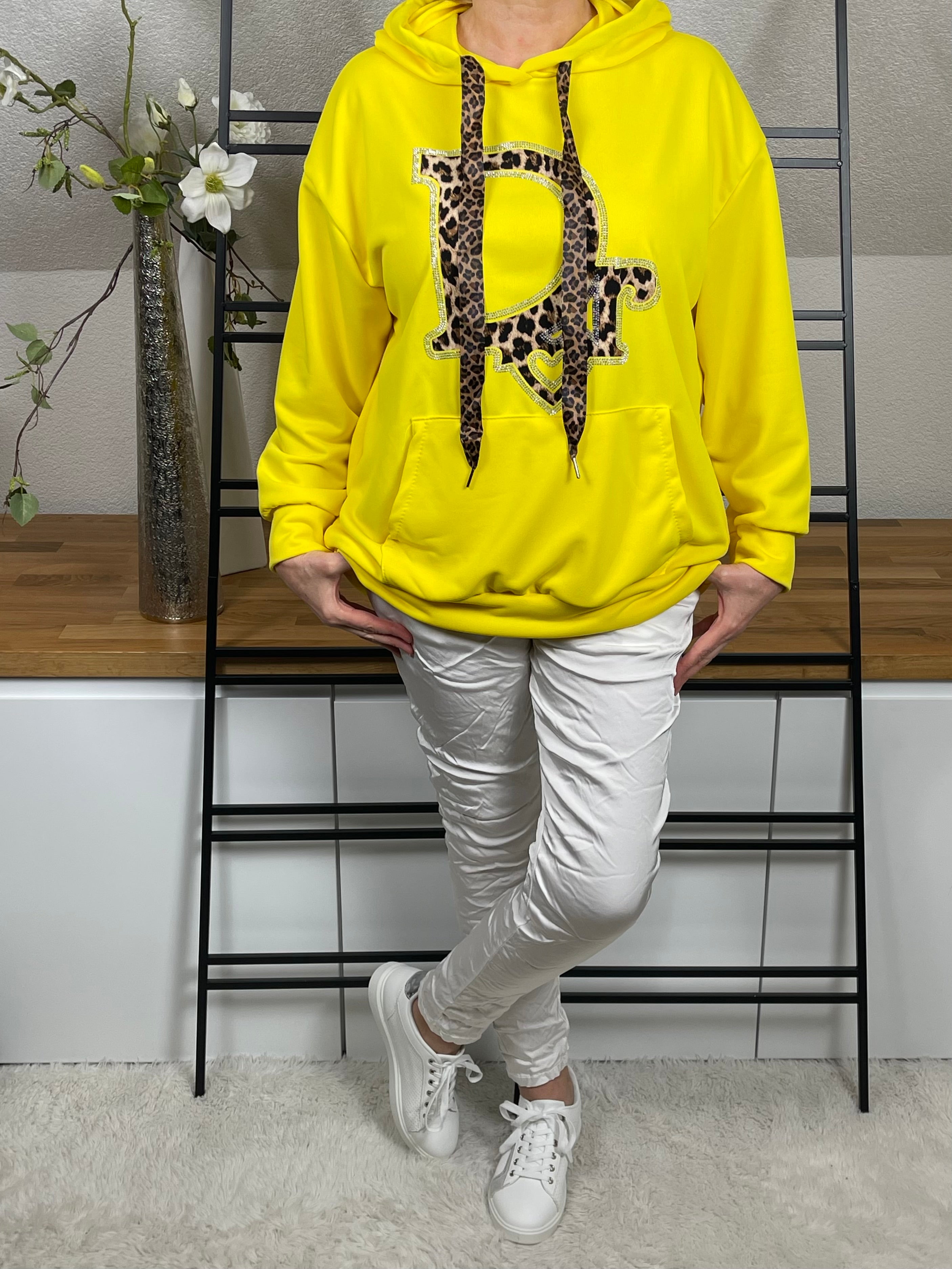 Frontansicht eines gelben Hoodies mit Animal-Print und Glitzerdetails, kombiniert mit weißer Shiney - Hose und Sneakern, mit gekreuzten Beinen 